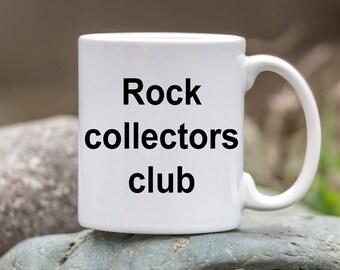 May include: White ceramic mug with black text that reads "Rock collectors club". The mug is sitting on a pile of gray rocks.