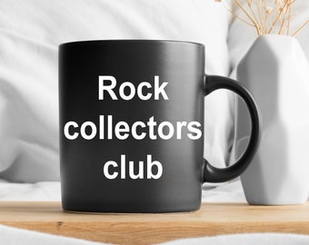 May include: Black ceramic mug with white text that reads "Rock collectors club" on a wooden tray. The mug is sitting on a white bedspread.