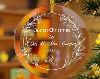 Pode incluir: Uma bola de Natal redonda e transparente com uma fita dourada. O enfeite apresenta desenhos florais brancos e o texto "Our 1st Christmas As Mr & Mrs Cooper 2022" com duas alianças. A bola está pendurada num galho verde.