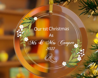 Pode incluir: Um enfeite de Natal de vidro transparente com uma fita dourada e um design floral branco. O enfeite diz "Our 1st Christmas As Mr & Mrs Cooper 2022" com dois anéis no centro.