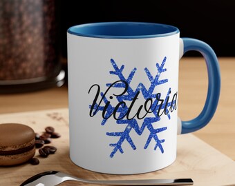 May include: White ceramic mug with a blue interior and handle. The mug features a blue glitter snowflake design and the name "Victoria" in a cursive font. The mug is on a wooden surface with a spoon and a pastry.
