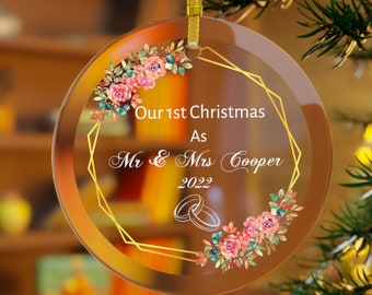 Pode incluir: Uma bola de Natal redonda e transparente com borda dourada e uma fita dourada para pendurar. O enfeite apresenta o texto "Our 1st Christmas As Mr & Mrs Cooper 2022" e detalhes florais em rosa, azul e verde.