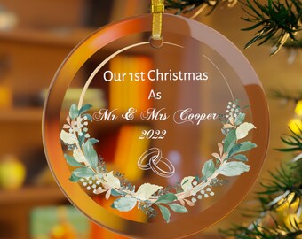 Pode incluir: Uma bola de Natal de vidro transparente com acabamento dourado e uma fita dourada para pendurar. O enfeite apresenta o texto "Our 1st Christmas As Mr & Mrs Cooper 2022" com uma coroa de flores e duas alianças.