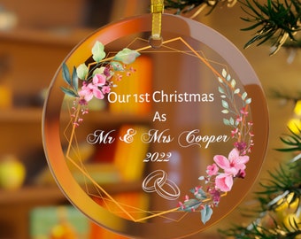Pode incluir: Enfeite de Natal de vidro transparente com borda dourada e fita dourada. O enfeite tem o texto "Our 1st Christmas As Mr & Mrs Cooper 2022" com detalhes florais e anéis. Está pendurado em uma árvore de Natal.