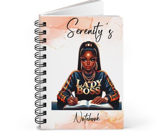May include: A spiral-bound notebook with a cover illustration of a woman in a black and gold sweatshirt. The sweatshirt reads "LADY BOSS". The notebook is titled "Serenity's Notebook" in a decorative script font, with a peach and gold background.