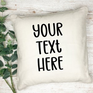 K&ouml;nnte beinhalten: Ein wei&szlig;es Kissen mit schwarzem Text, der "YOUR TEXT HERE" lautet. Das Kissen liegt auf einer wei&szlig;en Holzoberfl&auml;che.