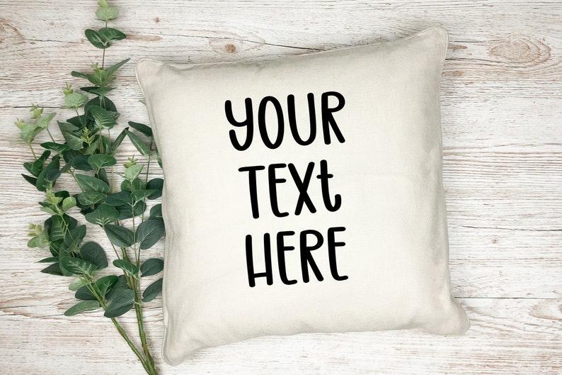 K&ouml;nnte beinhalten: Ein wei&szlig;es Kissen mit schwarzem Text, der "YOUR TEXT HERE" lautet. Das Kissen liegt auf einer wei&szlig;en Holzoberfl&auml;che.