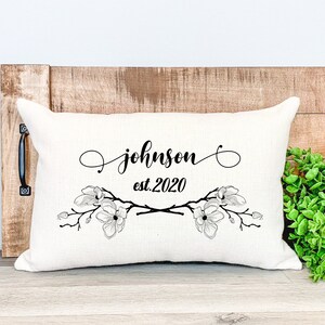 Könnte beinhalten: Cremefarbenes rechteckiges Kissen mit dem Namen "Johnson" und "est.2020" in schwarzer Schrift. Das Kissen hat ein Blumendesign mit Magnolienblüten. Das Kissen wird auf einer Holzoberfläche präsentiert.