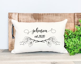 Könnte beinhalten: Cremefarbenes rechteckiges Kissen mit dem Namen "Johnson" und "est.2020" in schwarzer Schrift. Das Kissen hat ein Blumendesign mit Magnolienblüten. Das Kissen wird auf einer Holzoberfläche präsentiert.