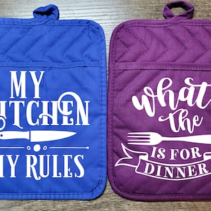 May include: Two quilted pot holders, one blue with the text "My Kitchen My Rules" and a knife graphic, and one purple with the text "What the is for Dinner" and a fork graphic.