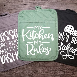 May include: Three kitchen pot holders with different sayings. The first is gray with the text "BLESS! are THOS WHO DO M DISH". The second is green with the text "-MY- Kitchen -MY- Rules". The third is black with the text "let's get Baked" and two cookie images.