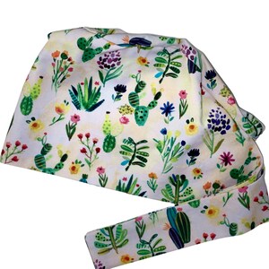 May include: A white scrub cap with a watercolour design of cacti, succulents, and wildflowers in green, yellow, pink, and purple. The cap has a tie closure for an adjustable fit.
