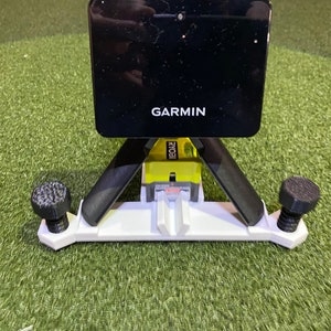 Könnte beinhalten: Ein schwarzes Garmin-Gerät ist auf einem weißen und gelben Stativständer montiert. Der Ständer hat schwarze Beine und schwarze Knöpfe. Auf dem Garmin-Gerät ist der Markenname in Weiß aufgedruckt. Der Ständer steht auf einer grünen Oberfläche.