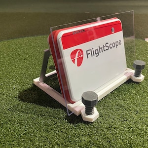 May include: A white and red FlightScope device with a clear plastic shield, mounted on a white and gray stand. The device is secured with gray bolts and sits on artificial turf. The FlightScope logo is visible.