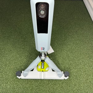 Puede incluir: Una cámara de seguridad gris claro con un sistema de doble lente, montada sobre una base de trípode. La base es blanca y gris con un acento amarillo y la marca "RYOBI". La cámara está sobre una superficie verde.