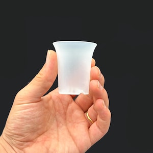 May include: A small, frosted glass cup with a flared rim is held in a hand. The cup is translucent, allowing light to pass through. The background is a solid black.