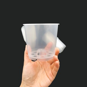 Puede incluir: Una maceta de plástico transparente con agujeros de drenaje en la parte inferior. La maceta se sostiene en una mano.