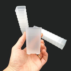 May include: A hand holds a clear, rectangular plastic container. Several more containers are stacked in the background. The containers are translucent and have a square opening. The background is a solid black.