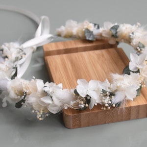 Puede incluir: Una corona de flores blancas con hojas verdes y plumas blancas. La corona est&aacute; sobre un soporte de madera.