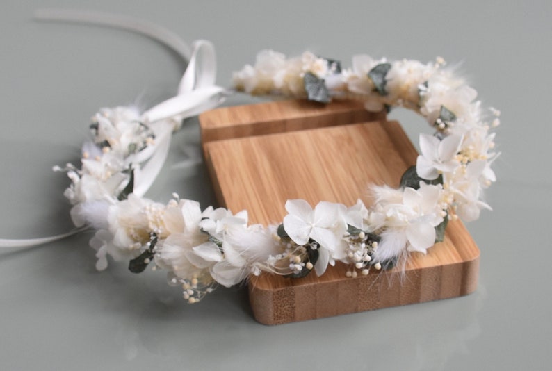 Puede incluir: Una corona de flores blancas con hojas verdes y plumas blancas. La corona est&aacute; sobre un soporte de madera.