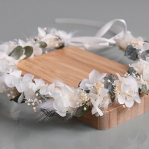 Puede incluir: Corona de flores blancas con hojas de eucalipto y plumas blancas. La corona est&aacute; atada con una cinta blanca y se muestra sobre una superficie de madera.