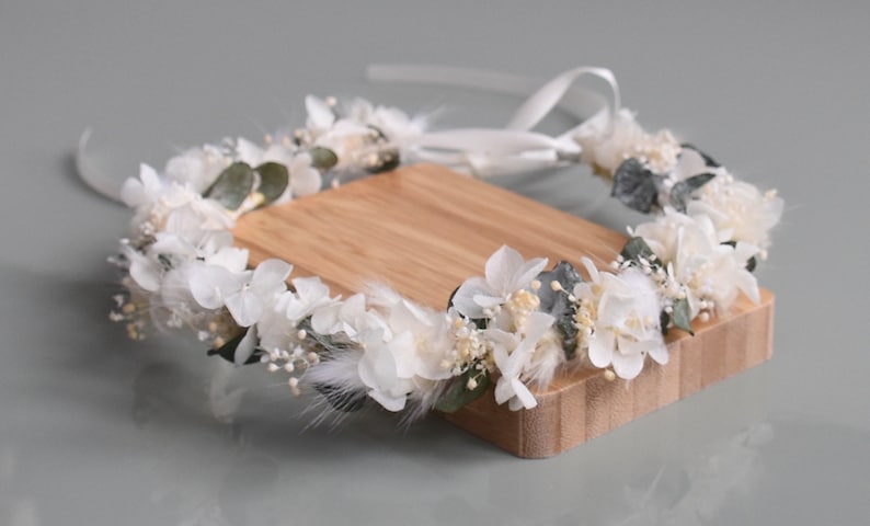 Puede incluir: Corona de flores blancas con hojas de eucalipto y plumas blancas. La corona est&aacute; atada con una cinta blanca y se muestra sobre una superficie de madera.