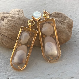 Könnte beinhalten: Goldfarbene Ohrringe mit rechteckigem Design, die weiße Muscheln in klarem Harz enthalten.