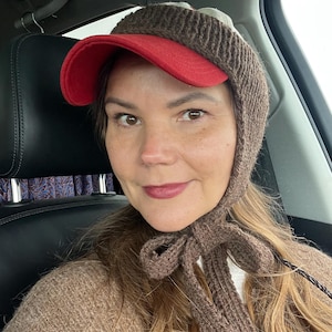 May include: A brown knitted bonnet with a red brim and a tie closure under the chin. The bonnet features a ribbed texture and covers the top and sides of the head, while the red brim extends forward from the forehead area.