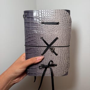 May include: A rectangular journal with a grey and light grey crocodile-embossed cover. The front features black ribbon lacing through silver metal eyelets, a thin black elastic band closure, and a small silver heart-shaped charm attached to the lace.