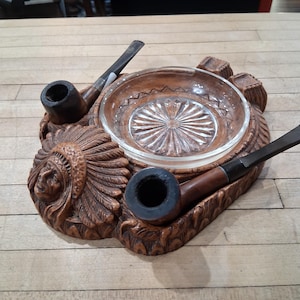 May include: A brown, carved wooden ashtray with a Native American chief design, featuring a central glass dish and two tobacco pipes. The ashtray is designed to hold two pipes and has a detailed, textured surface.