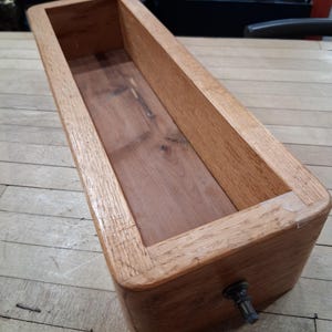 May include: A rectangular wooden planter box with a light brown finish. The box has a hollow interior and a dark metal spigot on one end. The wood grain is visible on the sides and bottom of the box. The planter is sitting on a wooden surface.