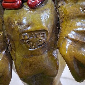 May include: Close-up of a weathered, olive-toned sculpture with a red and black accent. The sculpture has a textured surface and appears to be an antique or vintage piece. The details include a partial inscription, suggesting it may be a collectible item.