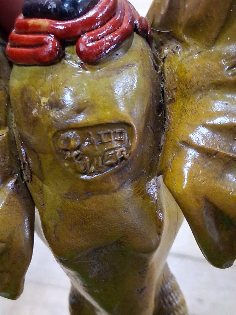 May include: Close-up of a weathered, olive-toned sculpture with a red and black accent. The sculpture has a textured surface and appears to be an antique or vintage piece. The details include a partial inscription, suggesting it may be a collectible item.