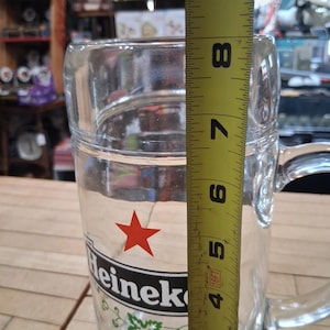 May include: A clear glass beer mug featuring the red Heineken star logo and the text Heineken Holland. A yellow measuring tape is positioned vertically against the side of the mug, indicating a height of approximately 8.5 inches from the base.