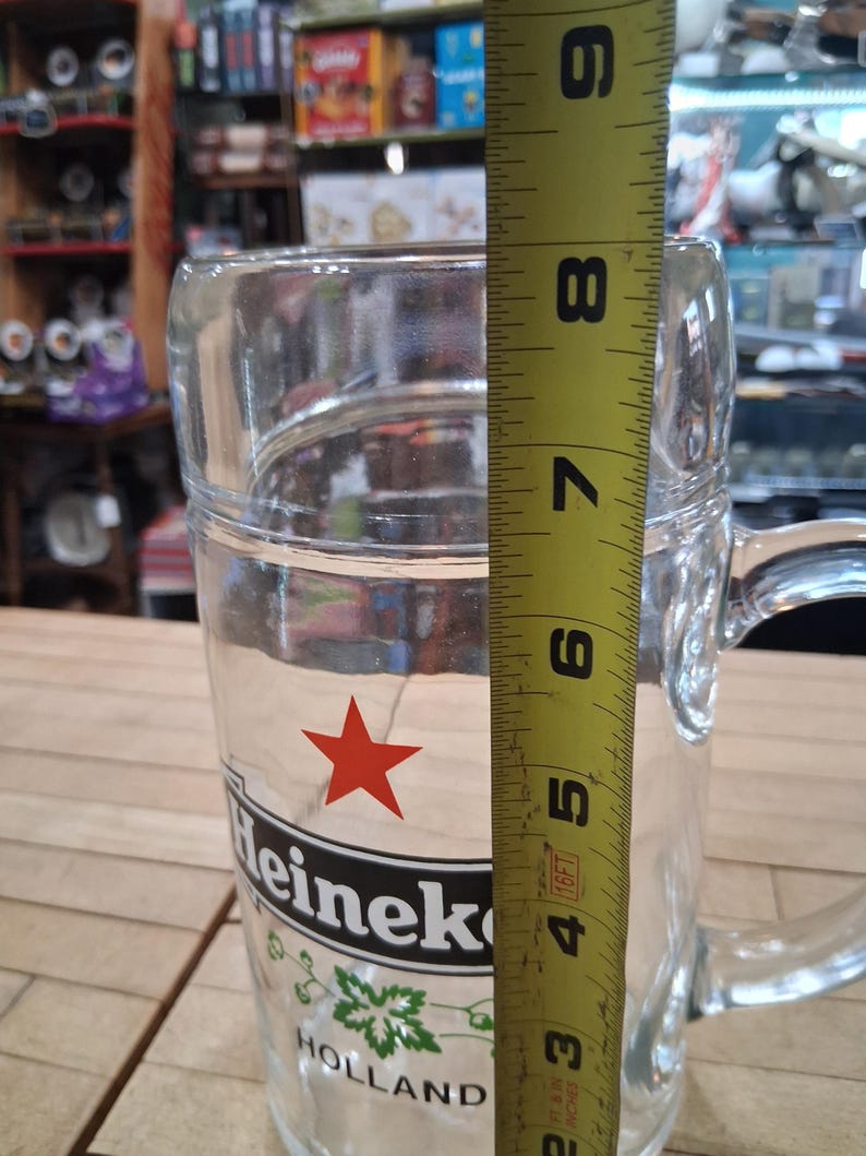 May include: A clear glass beer mug featuring the red Heineken star logo and the text Heineken Holland. A yellow measuring tape is positioned vertically against the side of the mug, indicating a height of approximately 8.5 inches from the base.