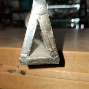 May include: A close-up of a silver metal object shaped like a triangle. The object has a dull, aged appearance with visible wear and tear. The edges are slightly rounded, and the object appears to be made of a solid metal.