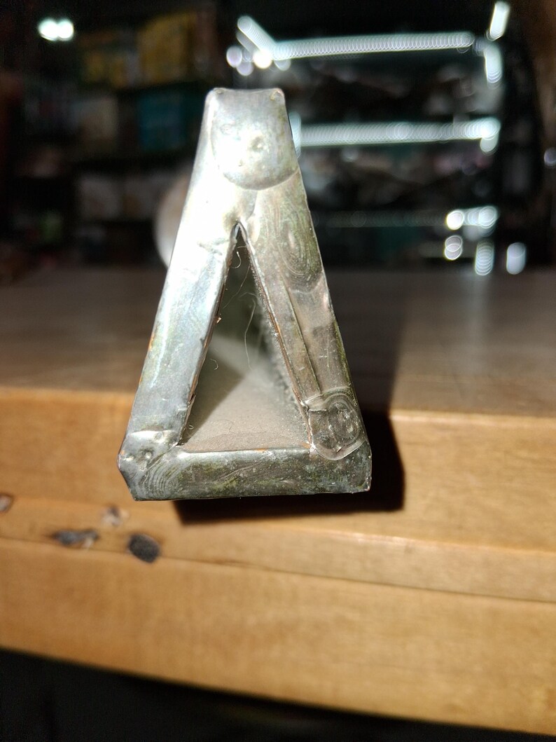 May include: A close-up of a silver metal object shaped like a triangle. The object has a dull, aged appearance with visible wear and tear. The edges are slightly rounded, and the object appears to be made of a solid metal.