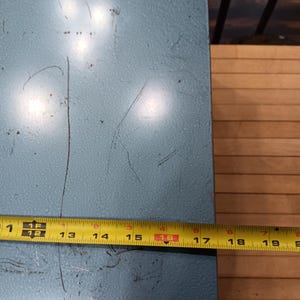 May include: A light blue metal surface with visible scratches. A yellow tape measure shows a length of approximately 16 inches. The surface appears to be part of a larger object, possibly a box or container.