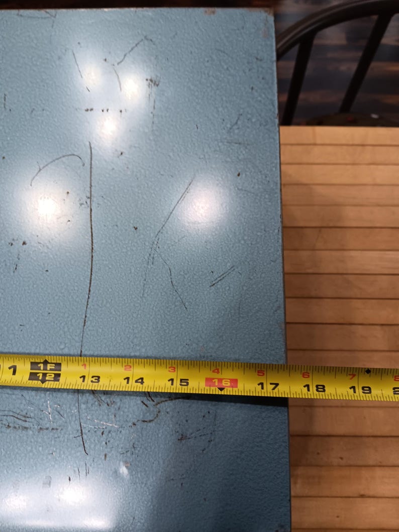 May include: A light blue metal surface with visible scratches. A yellow tape measure shows a length of approximately 16 inches. The surface appears to be part of a larger object, possibly a box or container.