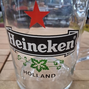 May include: A clear glass beer mug featuring a red star above a black banner with the white text Heineken. Below the banner, a green hop graphic is centered above the black text HOLLAND. The mug has a sturdy handle and a thick, circular base.