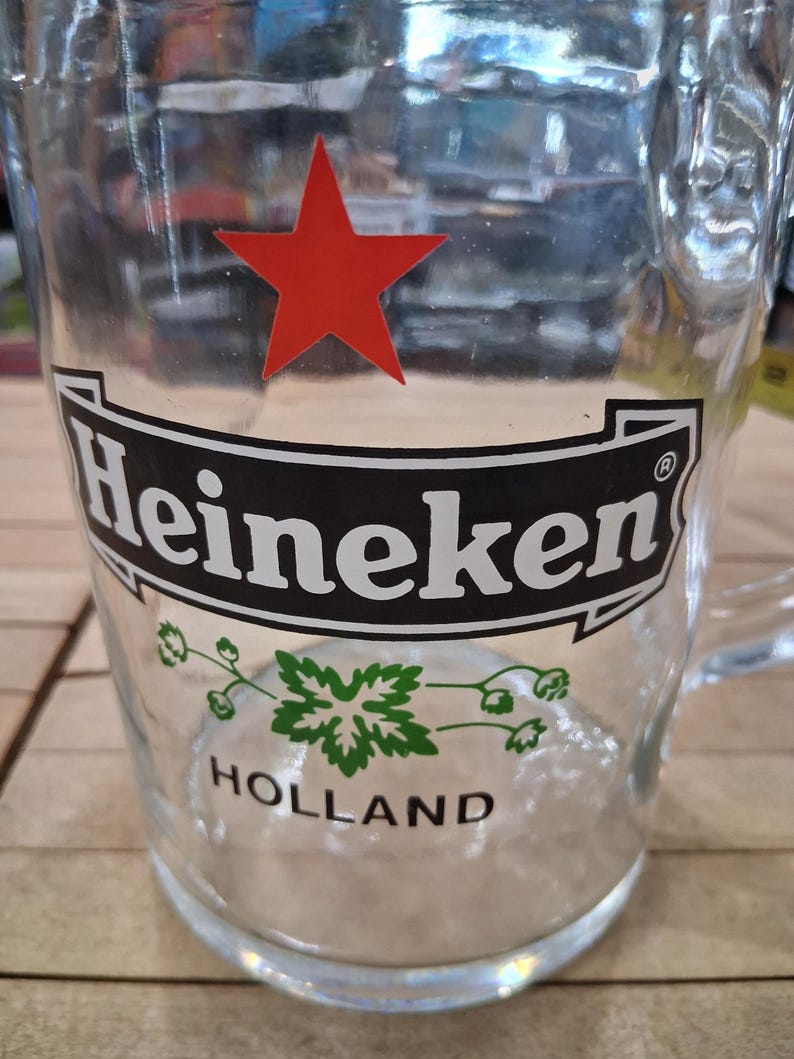 May include: A clear glass beer mug featuring a red star above a black banner with the white text Heineken. Below the banner, a green hop graphic is centered above the black text HOLLAND. The mug has a sturdy handle and a thick, circular base.