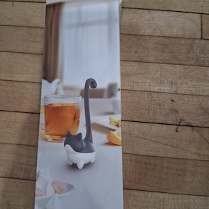 May include: A cat-shaped tea infuser, with a gray body and a white base, is featured on a product box. The infuser's tail extends upwards. A glass of tea and a lemon slice are visible in the background, suggesting tea time.