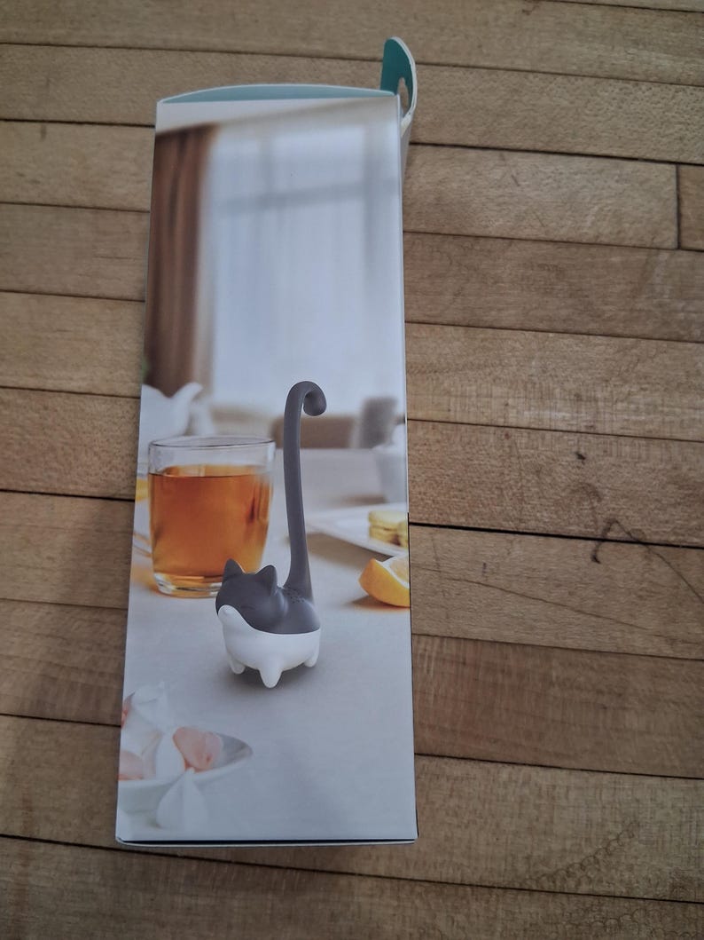 May include: A cat-shaped tea infuser, with a gray body and a white base, is featured on a product box. The infuser's tail extends upwards. A glass of tea and a lemon slice are visible in the background, suggesting tea time.