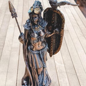 May include: A bronze-toned statue of a woman in classical attire, holding a spear and shield. An eagle with outstretched wings is perched on the shield. The statue has intricate details, including a decorative helmet and flowing robes. The statue is on a wooden surface.