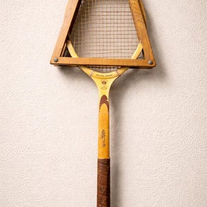 May include: Vintage wooden tennis racket with a brown leather grip. The racket features a wooden frame and a stringed net. The handle is yellow with text and a brown grip. The racket is a classic piece of sports equipment.