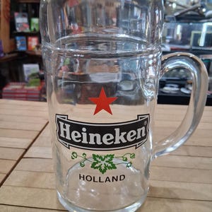 May include: A clear glass beer mug featuring a red star above the black and white Heineken logo. Below the logo is a green hop graphic and the word HOLLAND printed in black. The mug has a sturdy handle and a thick, cylindrical glass body.