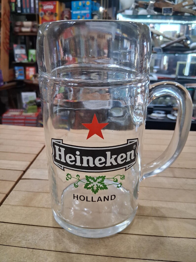 May include: A clear glass beer mug featuring a red star above the black and white Heineken logo. Below the logo is a green hop graphic and the word HOLLAND printed in black. The mug has a sturdy handle and a thick, cylindrical glass body.