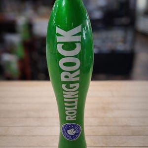 May include: A green Rolling Rock beer tap handle with white lettering. The tap handle has a silver top and bottom. A small blue and white emblem is near the bottom of the handle.