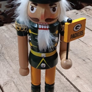 May include: Wooden Packers nutcracker figurine. The figurine is wearing a green and gold uniform and a tall hat with the Packers logo. It holds a sign that says "Packers". The base is green with a football.