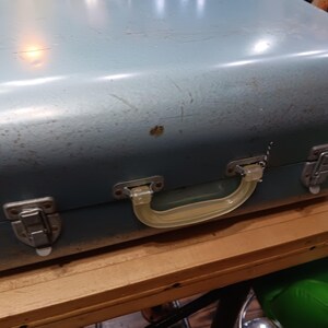 May include: A vintage, blue-green metal suitcase with a clear plastic handle and metal latches. The suitcase shows signs of wear, with scratches and a few spots of discoloration. The handle is a light yellow color.